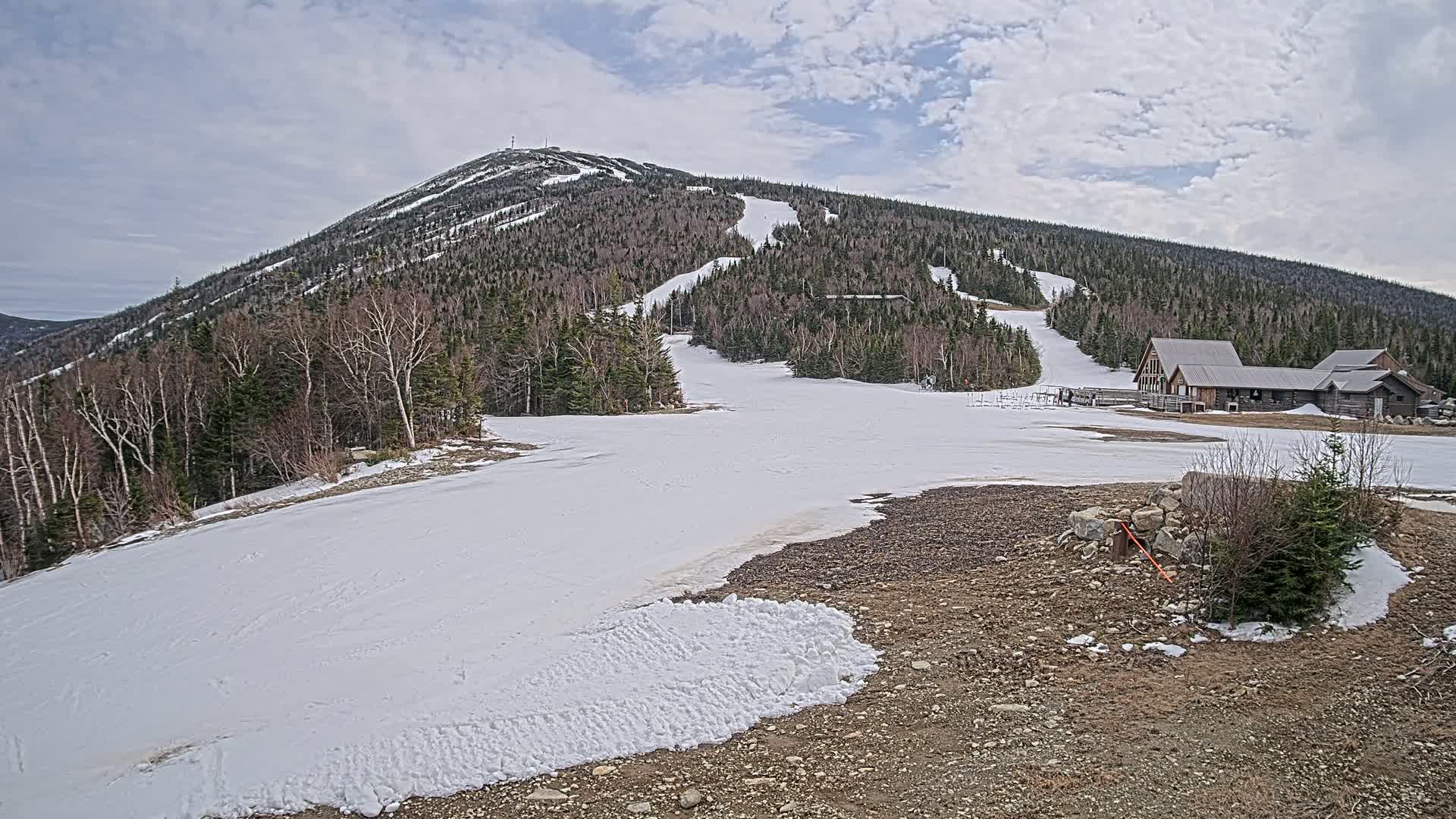 A snow-covered ski slope on a mountain with several buildings at the base under a cloudy sky.