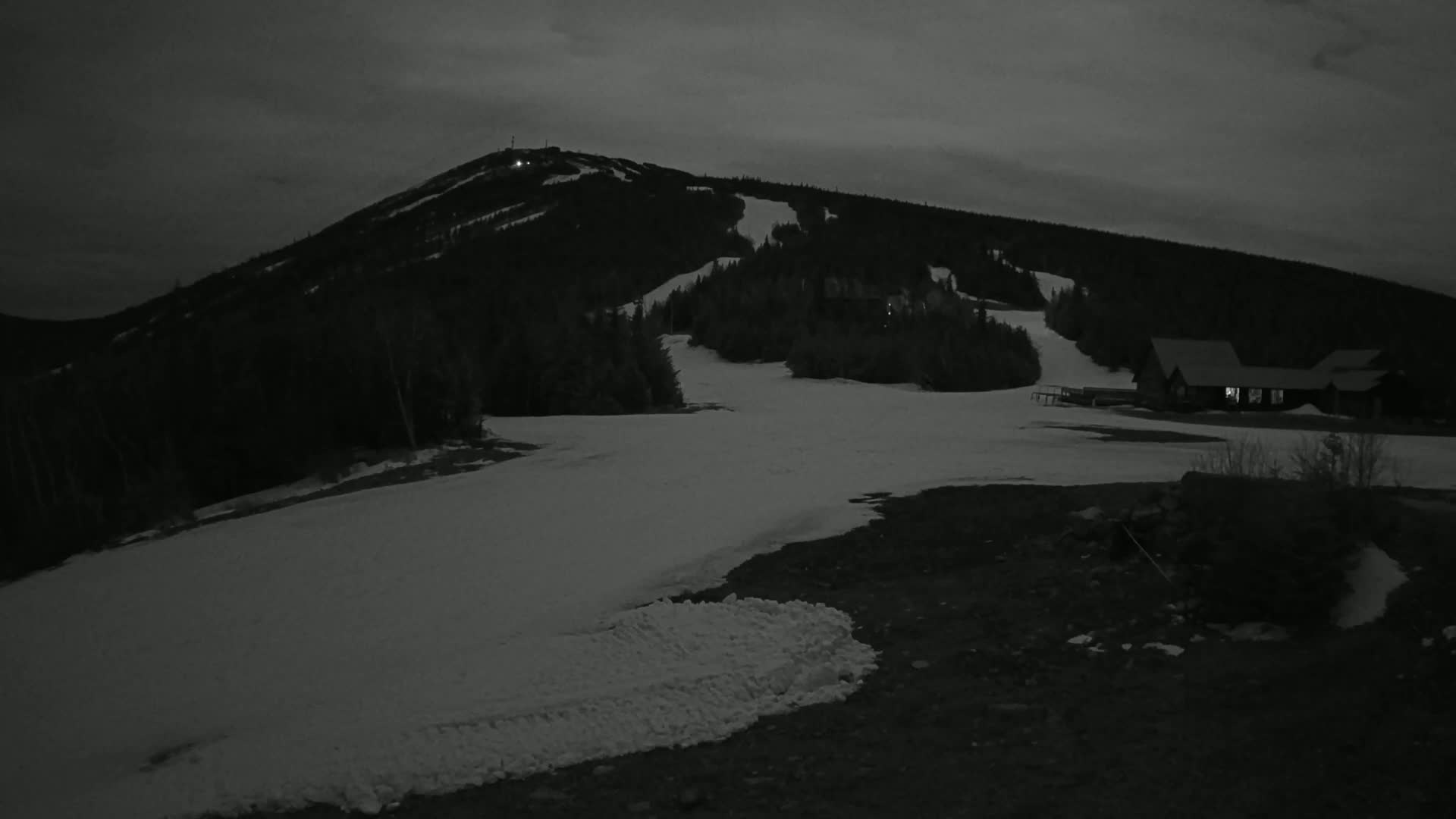 A snow-covered ski slope at night under a dark sky, with a building illuminated in the lower right and a mountain in the background.
