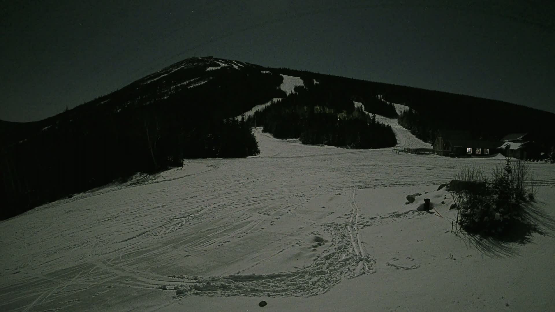 A snow-covered ski slope at night under a starry sky, with a mountain in the background and a building illuminated in the distance.