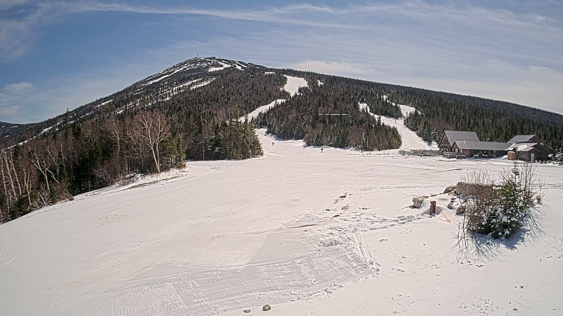 A snow-covered ski slope at the base of a mountain on a partly sunny day, with a few skiers visible and a ski lodge nearby.