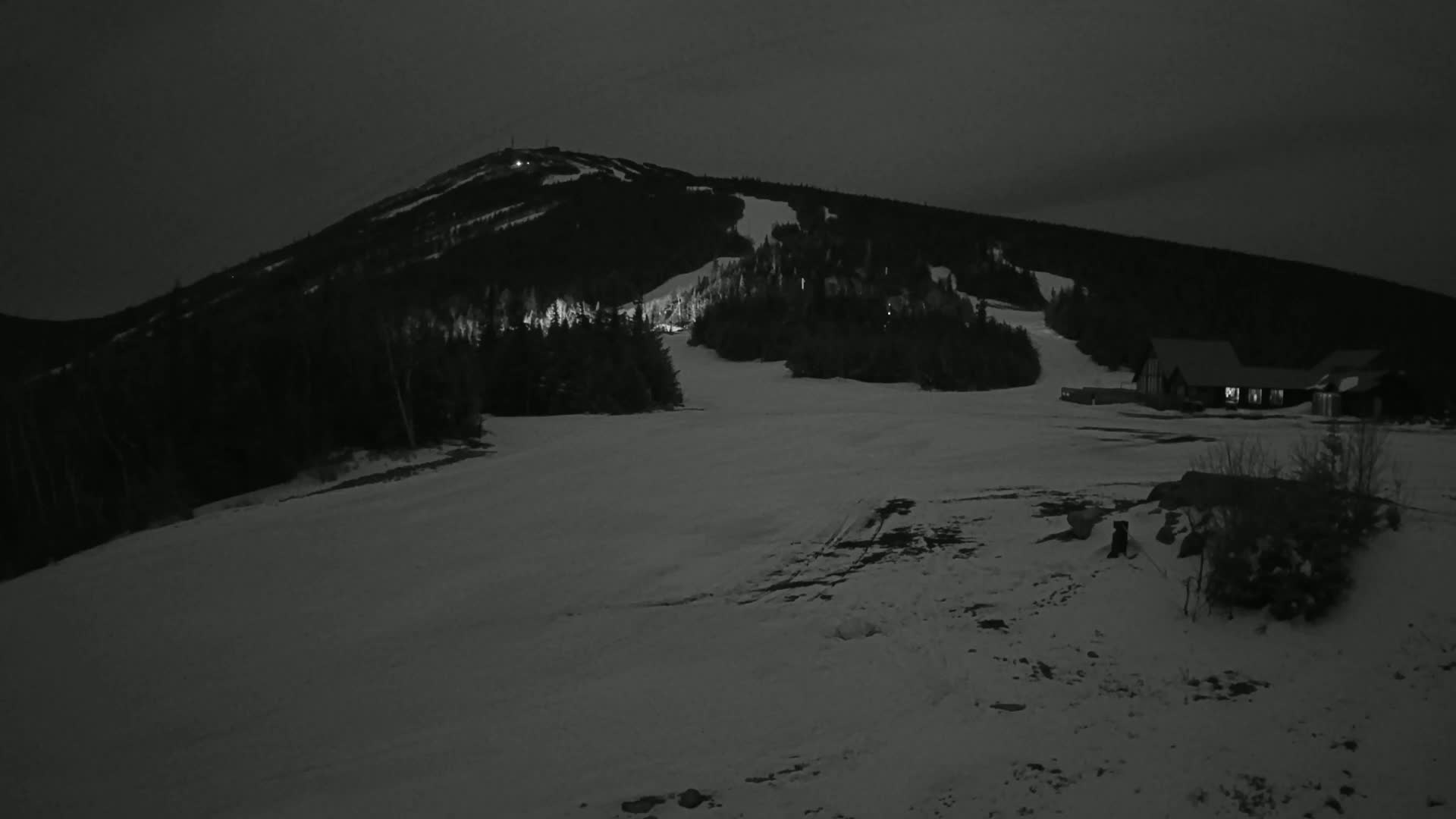 A snow-covered ski slope at night, with a mountain in the background and a building illuminated in the foreground.