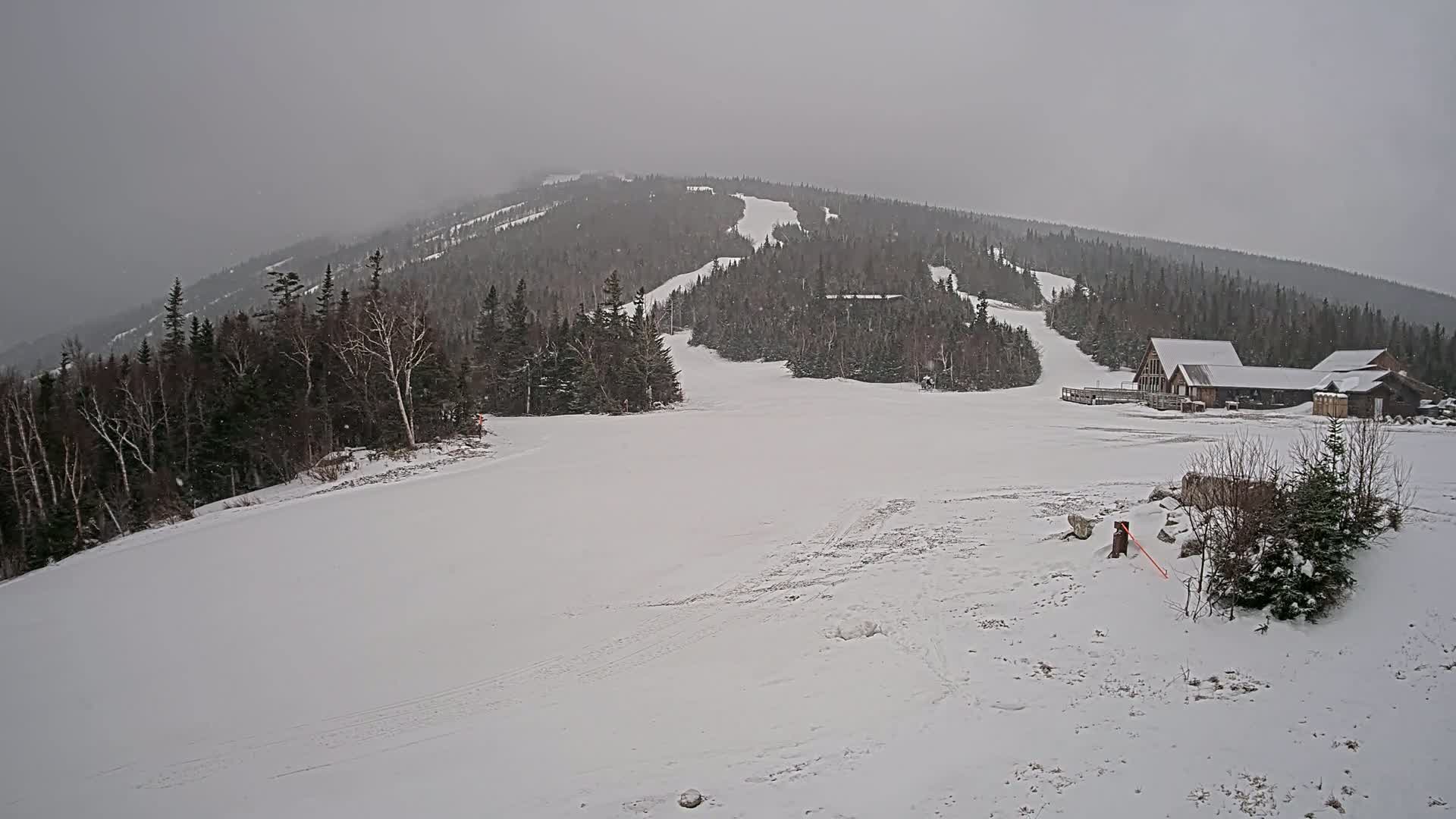 A snow-covered ski slope with a lodge at the base and a snow-covered mountain in the background under a gray, overcast sky.