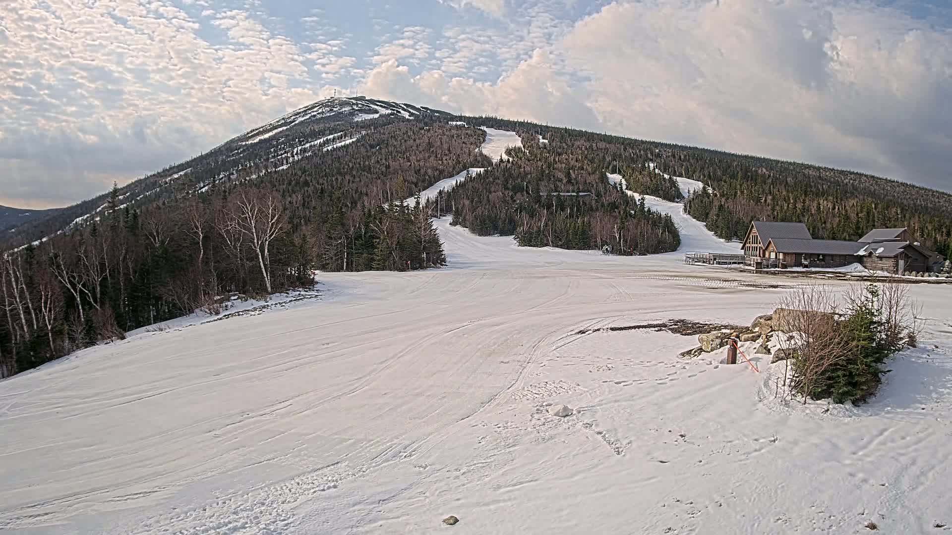 A snow-covered ski slope at the base of a mountain, under a partly cloudy sky, with a ski lodge visible in the background.