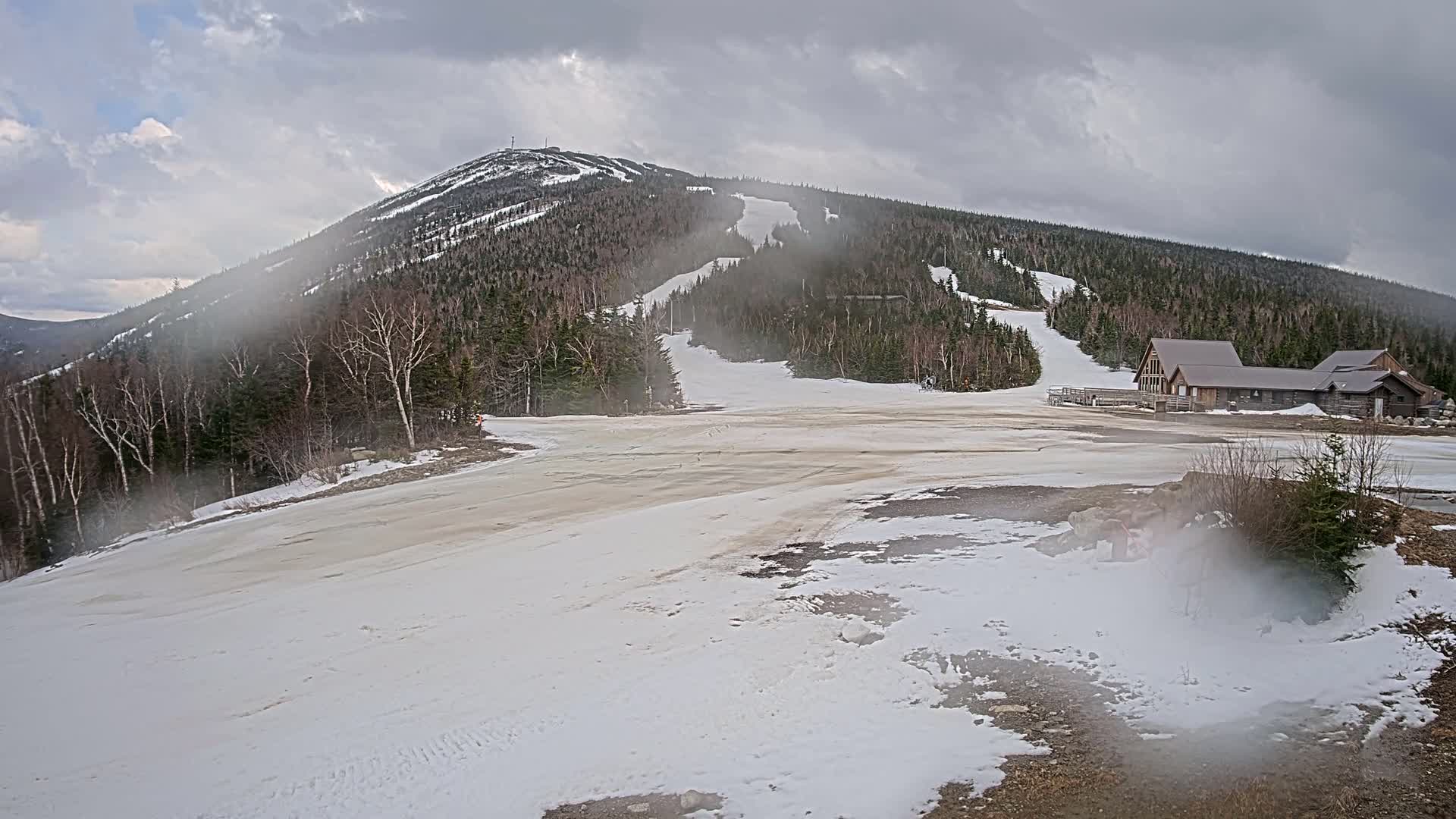 A mostly cloudy day shows a snow-covered ski resort with a mountain in the background, featuring a lodge at the base and partially melted snow on the lower slopes.