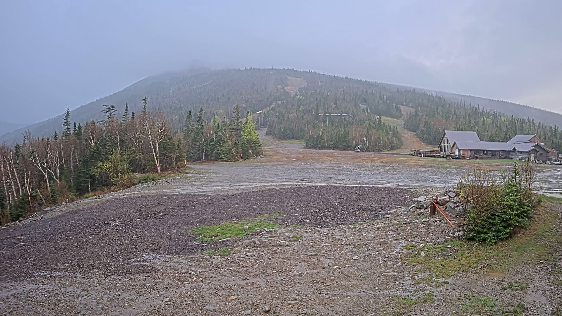 A rocky, muddy expanse in front of a ski lodge at the base of a forested mountain under an overcast sky.