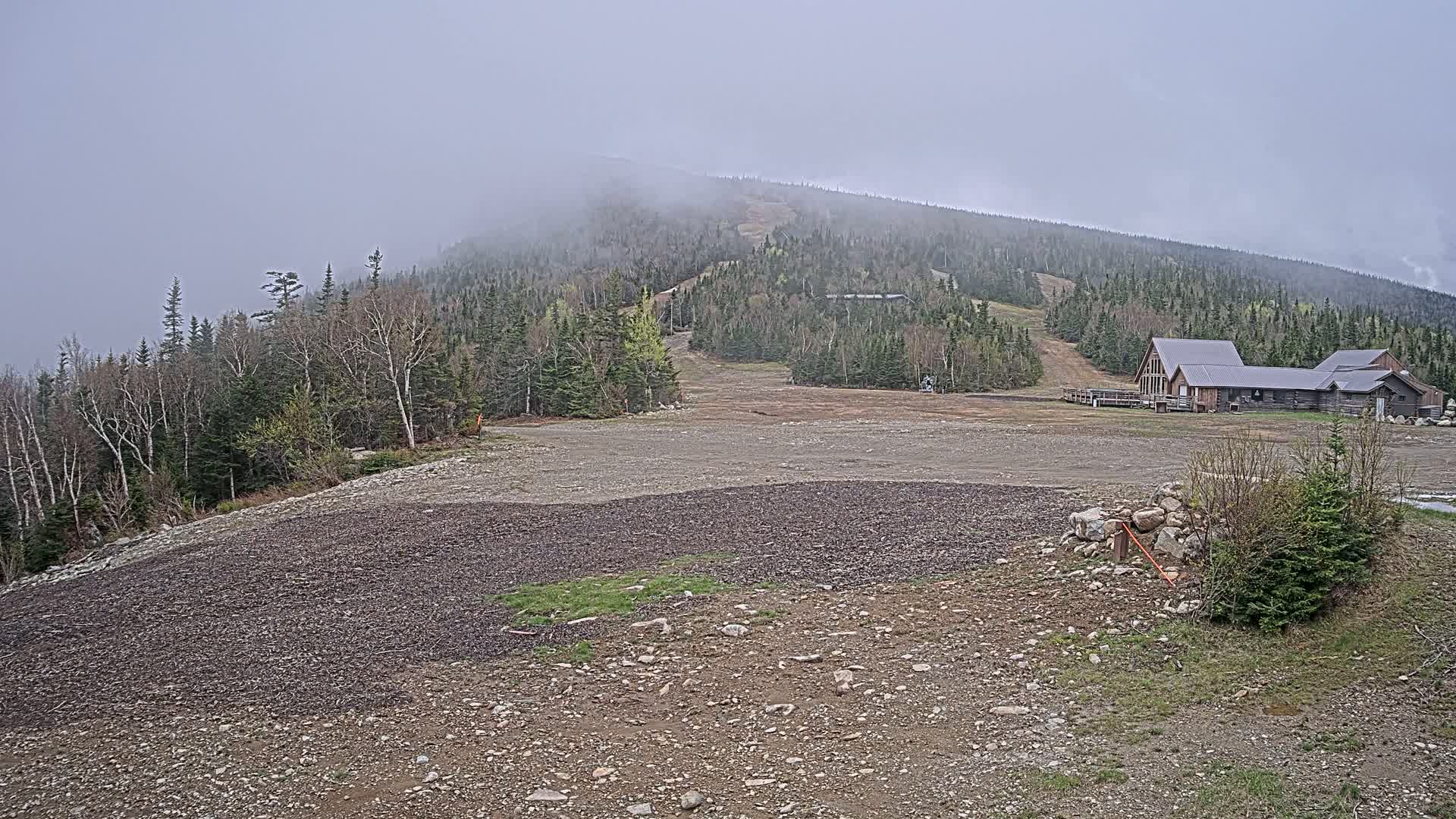 A rocky, barren landscape at the base of a fog-covered mountain reveals a ski slope and a long, low building.