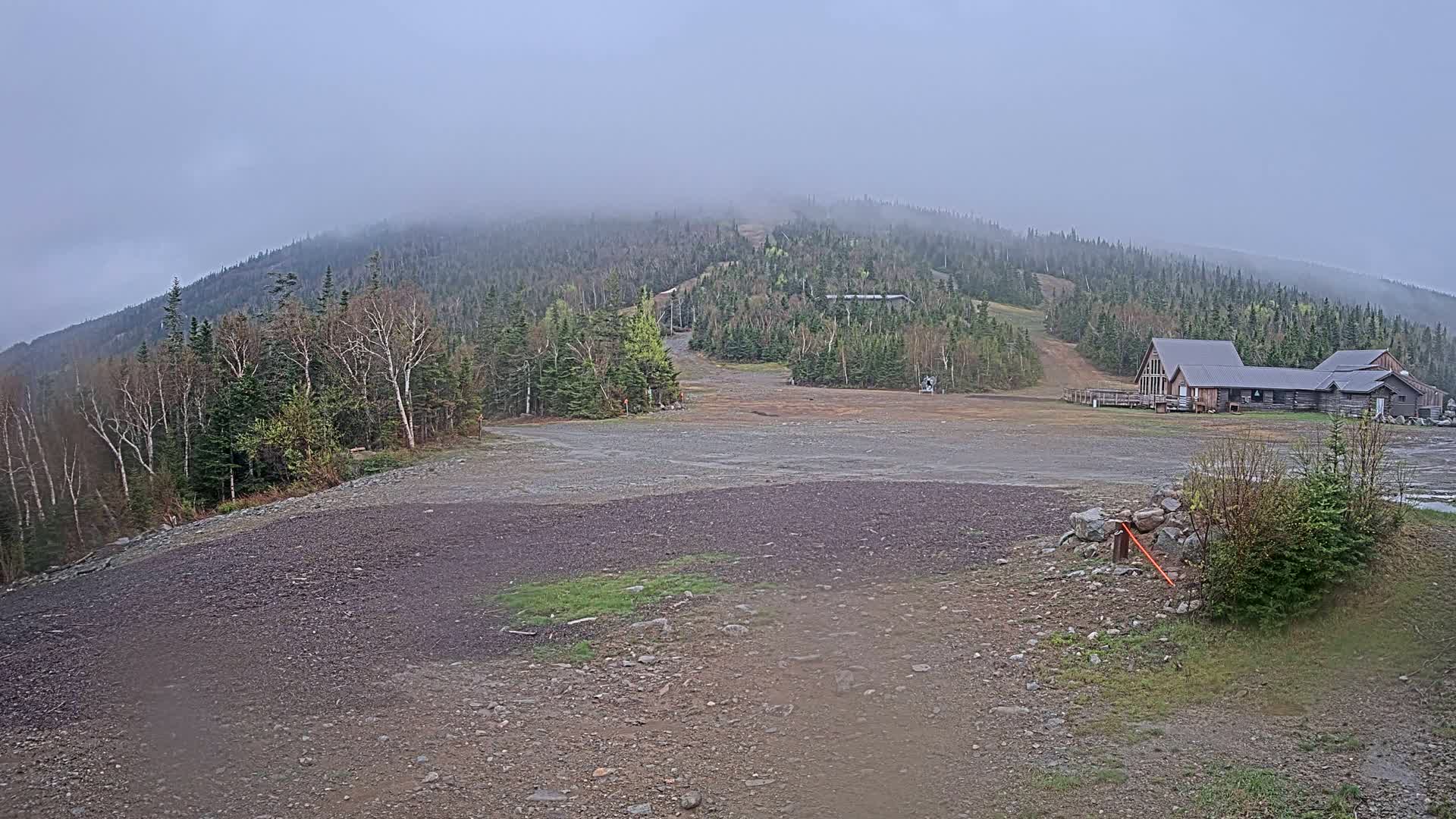 A mostly cloudy day reveals a ski resort with a large, rocky parking lot in the foreground and a wooden lodge nestled amongst a forested hillside.