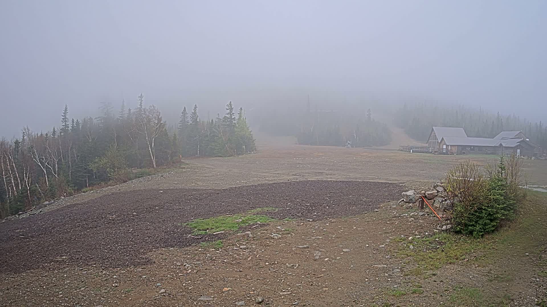 A foggy landscape shows a rocky, barren hillside sloping down toward a distant, low-lying building in a coniferous forest.