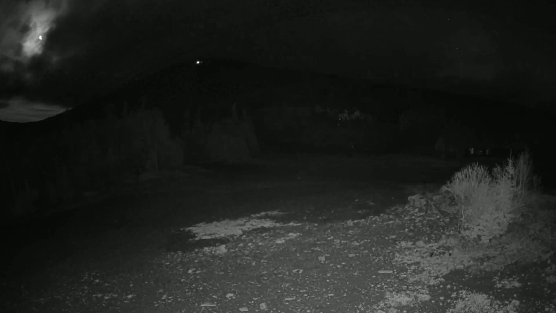 A dark, nighttime scene shows a rocky, gravelly ground in the foreground, with a hill silhouetted against a cloudy sky where a partially visible moon is present.