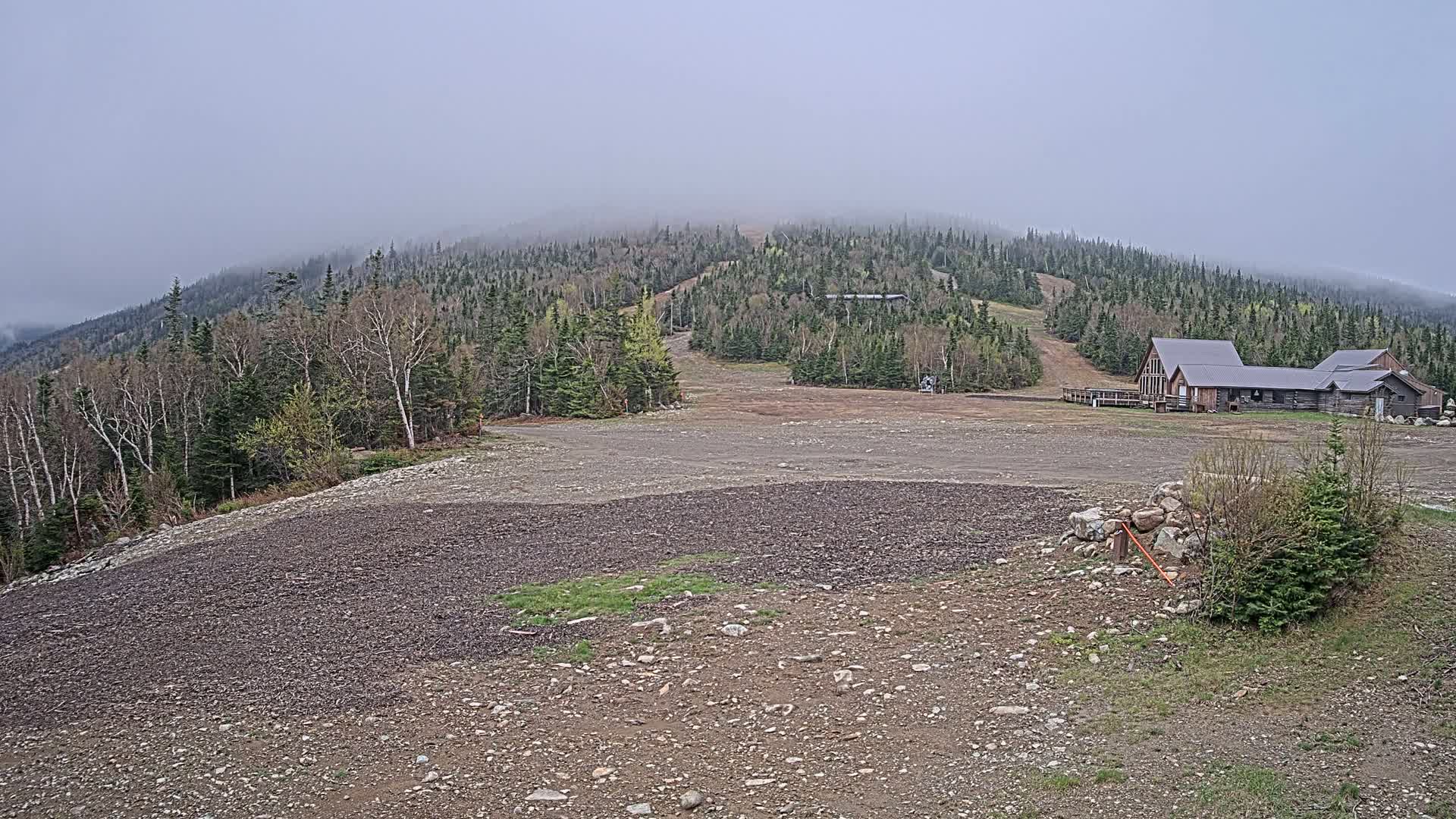 A gravel lot sits in front of a rustic lodge, overlooking a mostly barren ski slope on a foggy, overcast day.