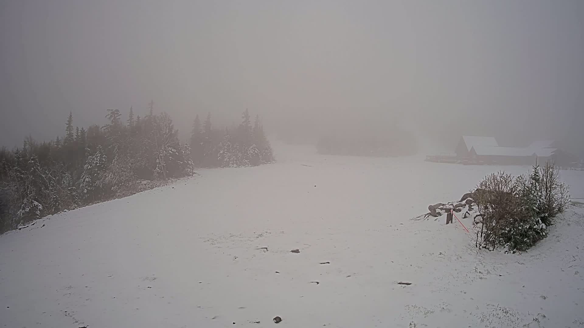 A snow-covered landscape with a few snow-covered buildings in the distance is shrouded in dense fog.