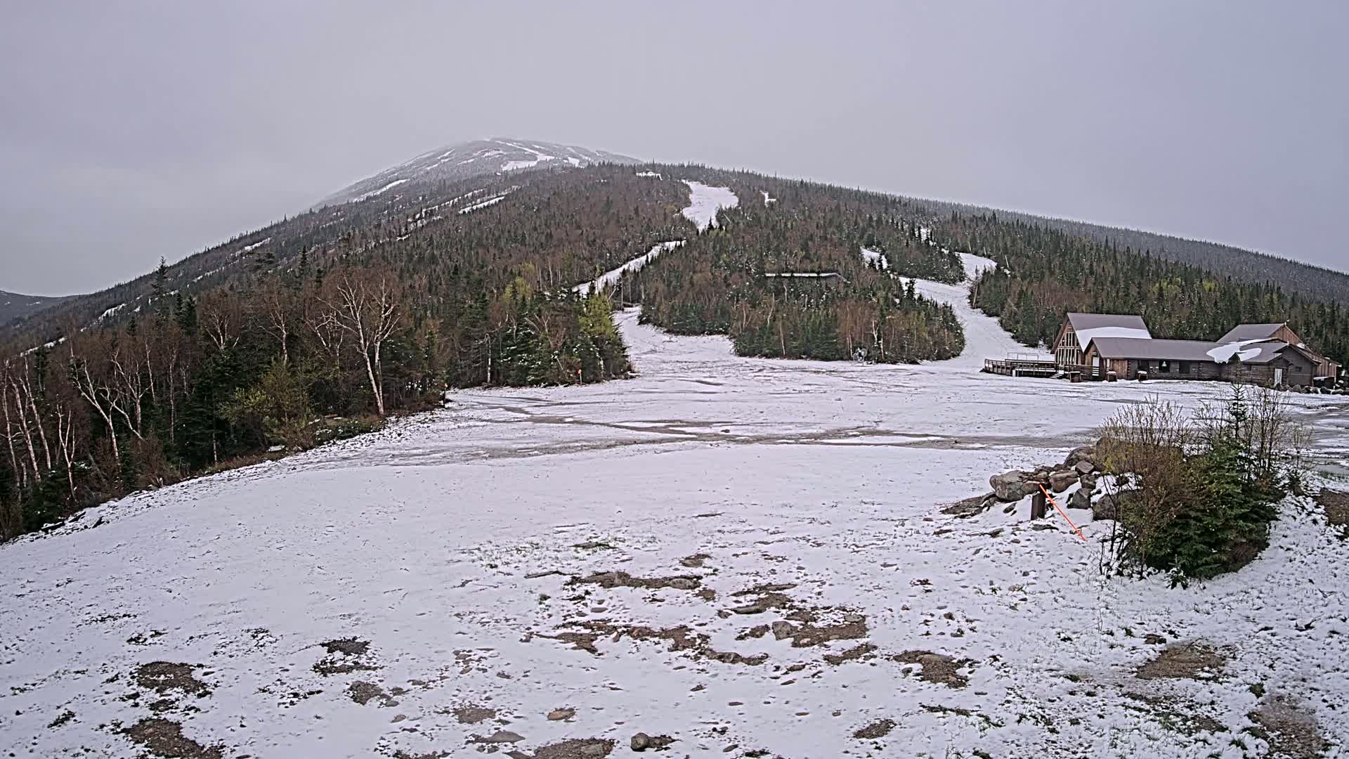 A snow-covered ski resort base area sits below a mountain with partially snow-covered ski trails, under an overcast sky.