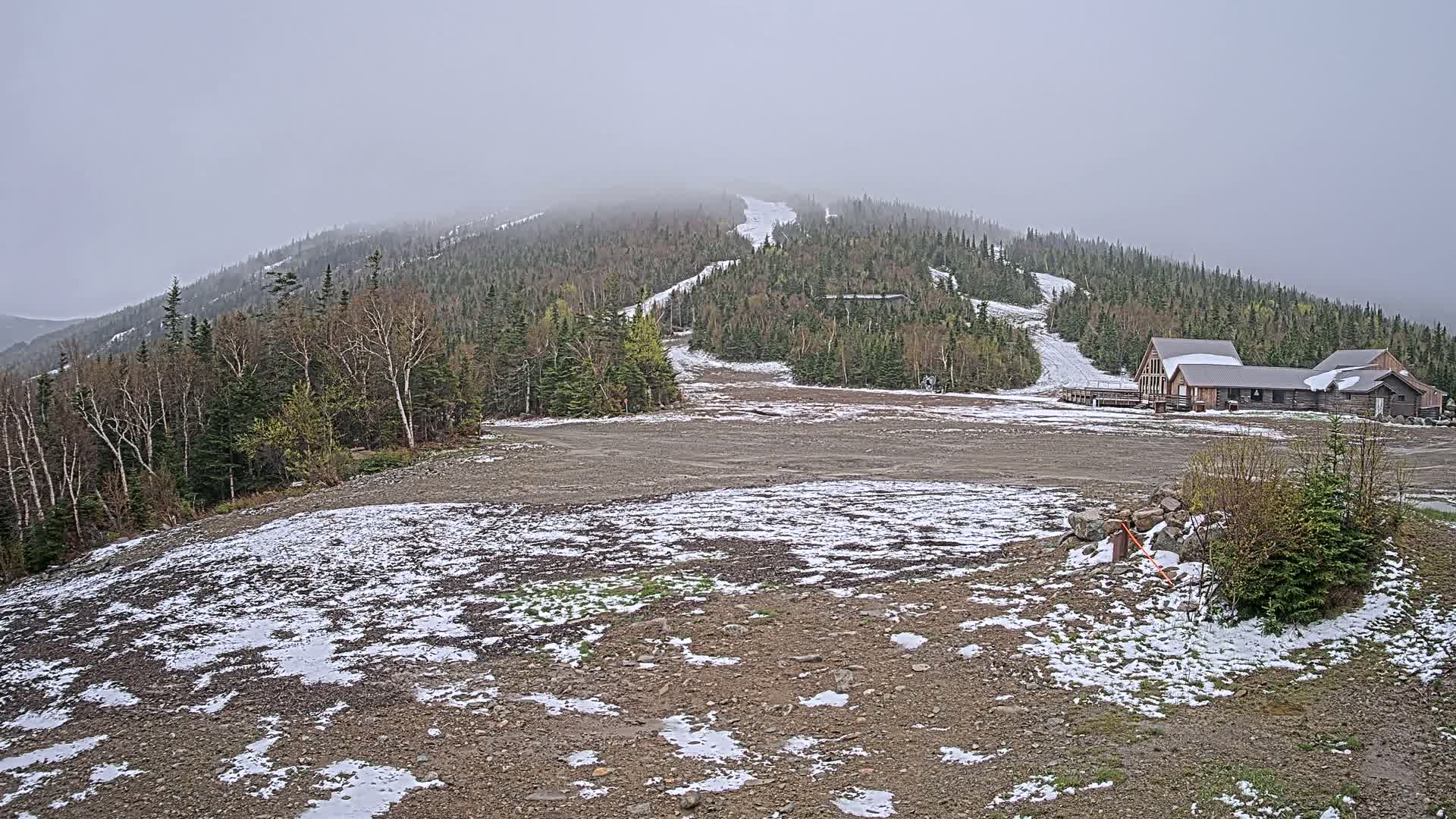 A mostly clear, snow-dusted ski resort base area with a wood lodge and a forested mountain under an overcast sky.
