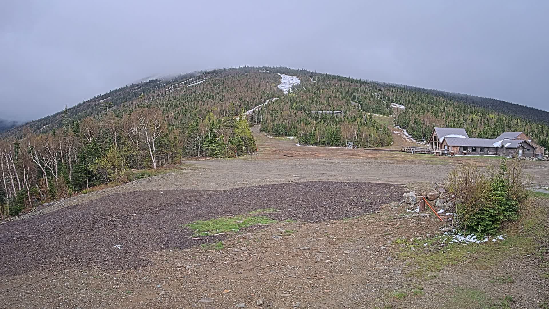 A mostly barren, rocky area with a few patches of grass and some lingering snow sits in front of a wood-framed building at the base of a forested mountain under an overcast sky.