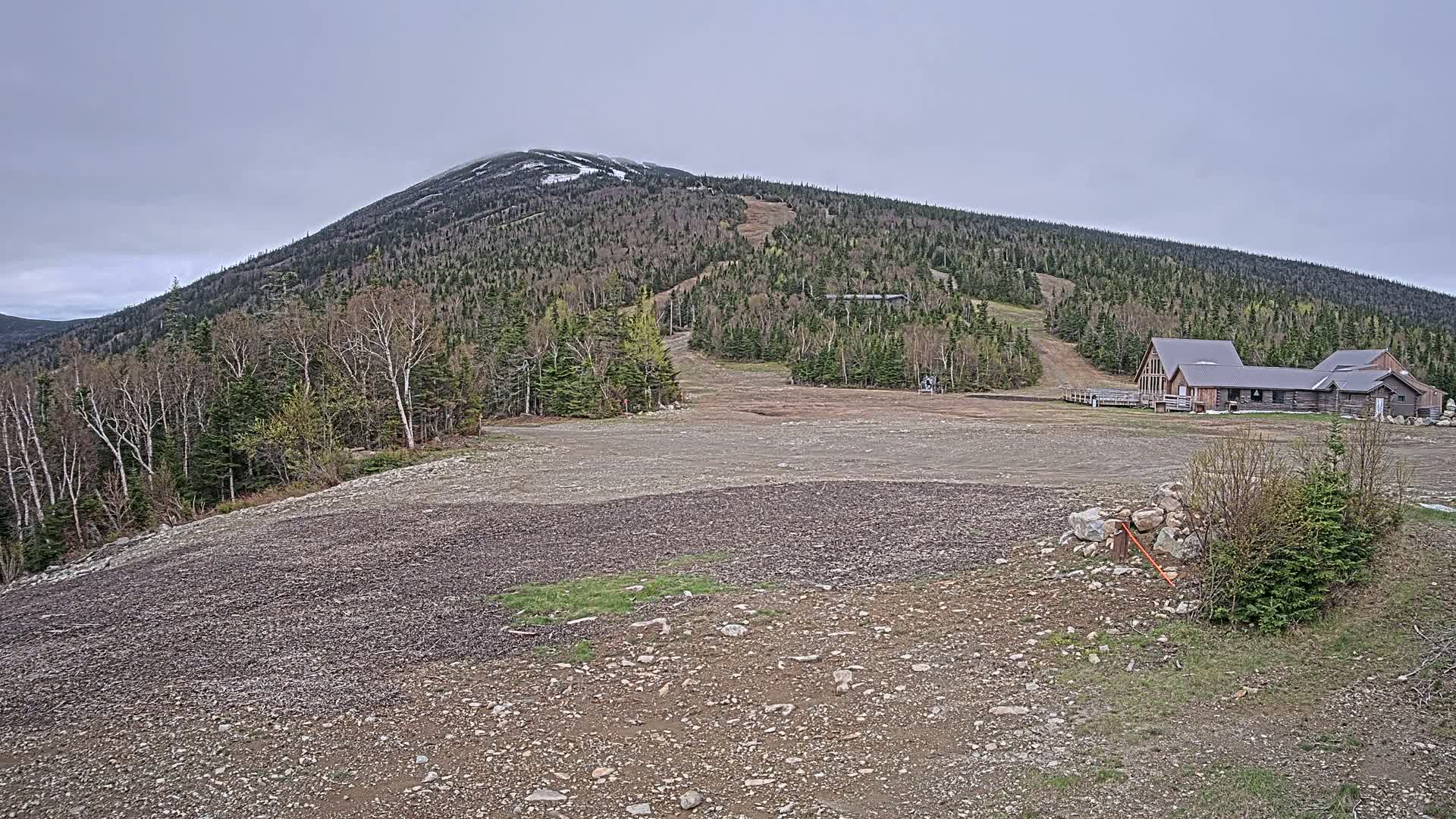 A rocky, barren landscape stretches toward a wooded mountain with a few patches of snow under an overcast sky, featuring a small lodge at the mountain's base.