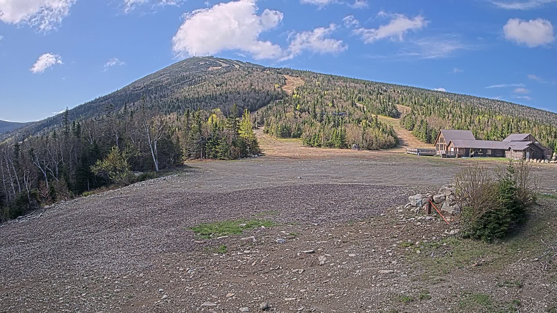 A rocky, mostly barren expanse stretches before a large, forested mountain with visible ski trails under a mostly sunny sky.