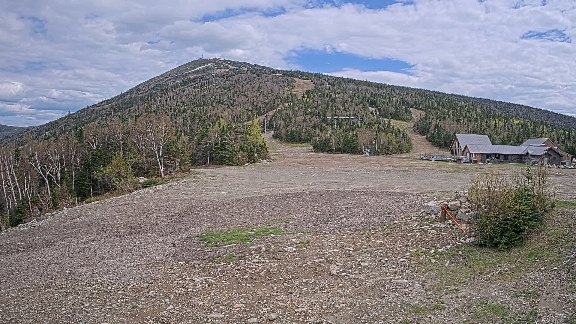 A rocky, gravelly area sits in front of a ski lodge at the base of a large, forested mountain under a partly cloudy sky.