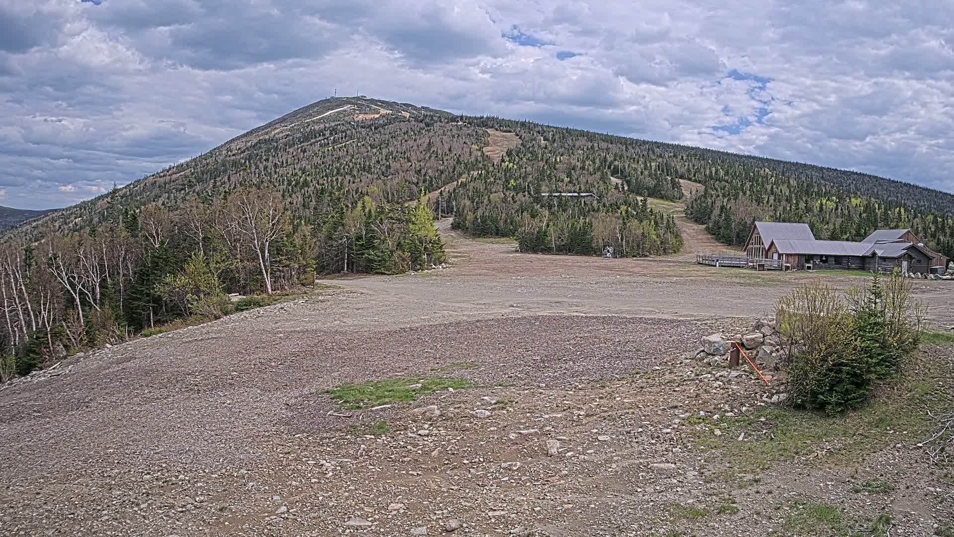 A rocky, empty expanse overlooks a wooded mountain with a ski resort building and cloudy skies.