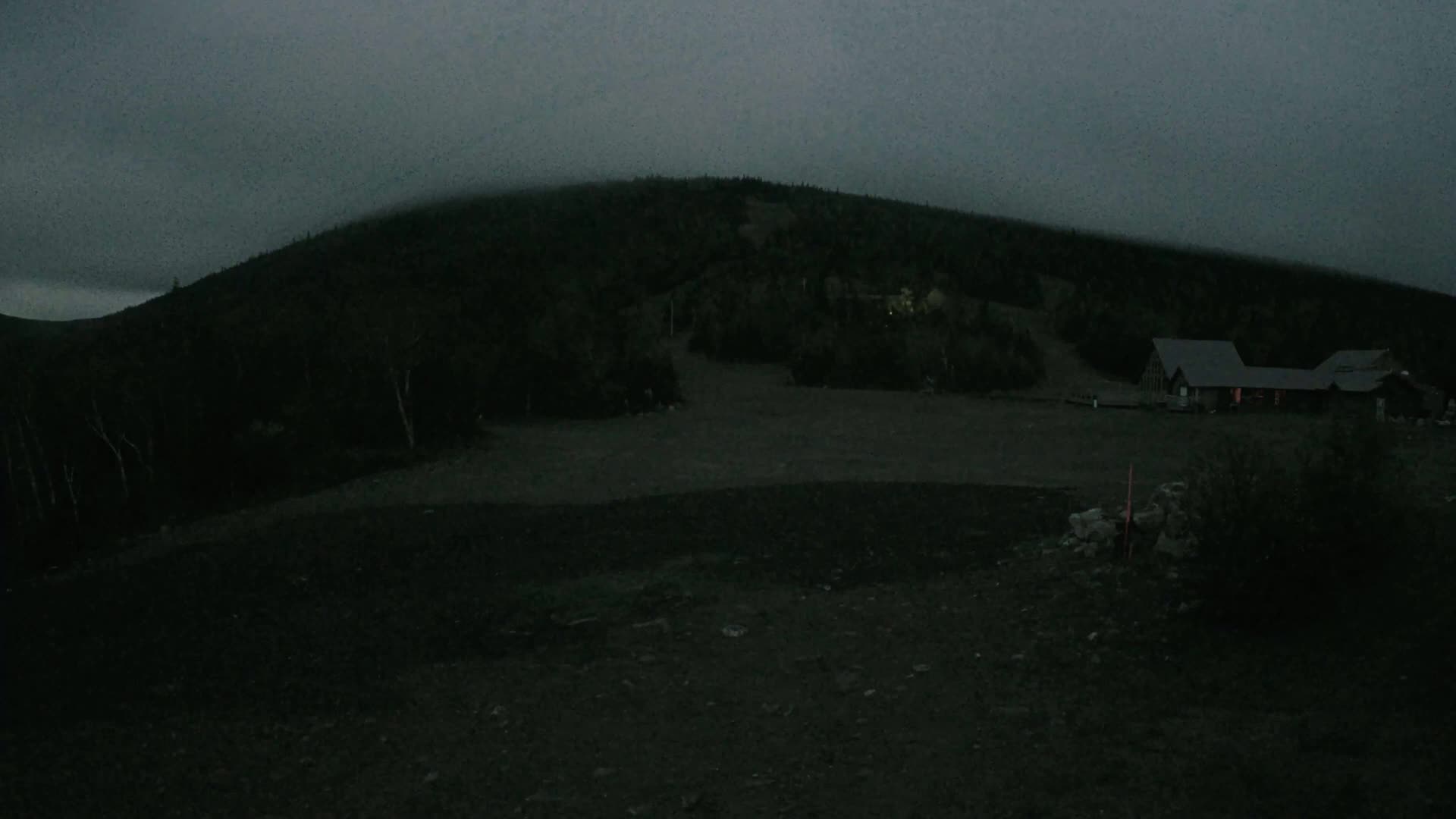 A dark, low-light image shows a building near the base of a hill covered in trees under an overcast sky.
