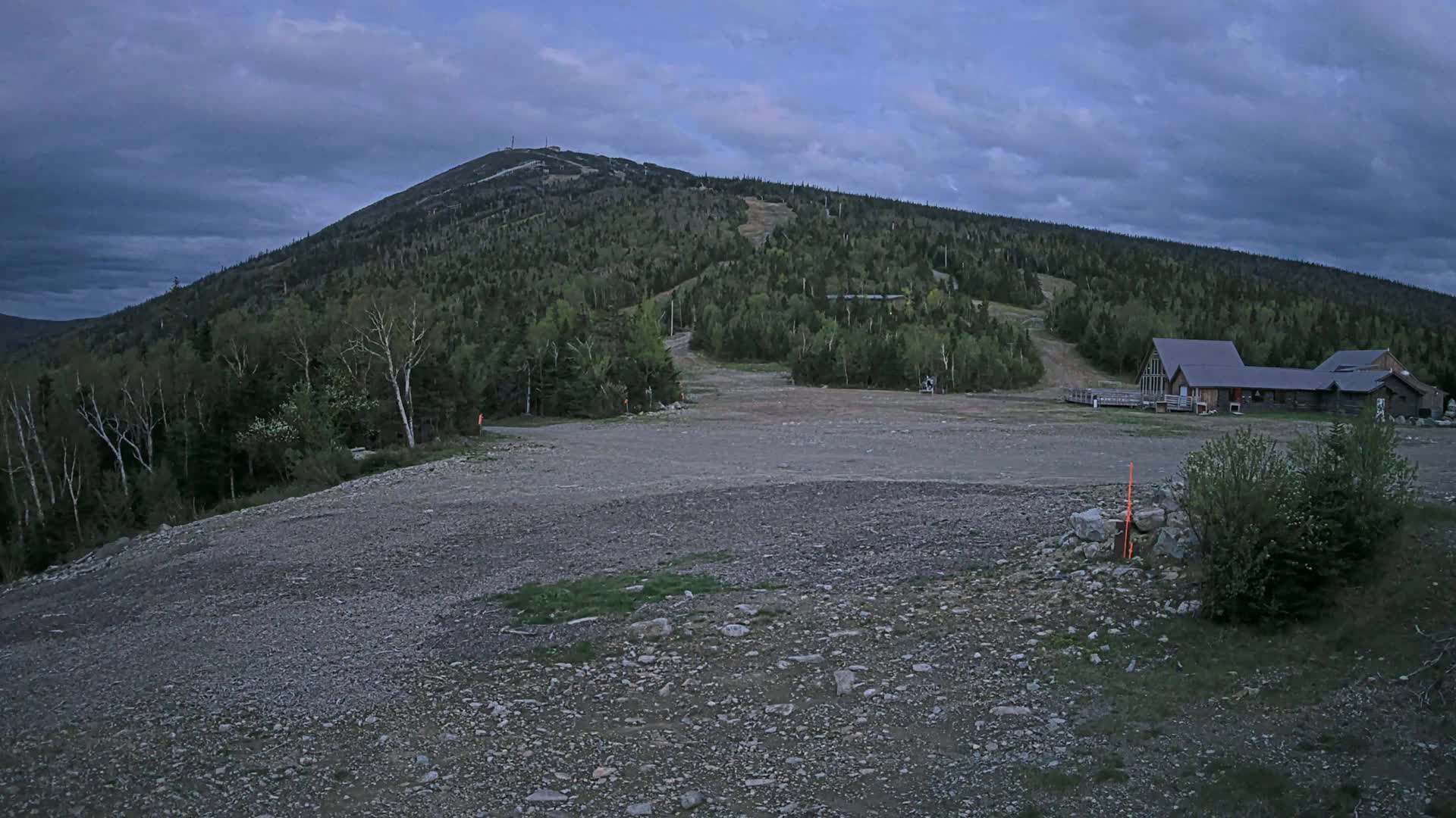 A rocky, empty area at the base of a tree-covered mountain under a cloudy sky sits before a small building complex.