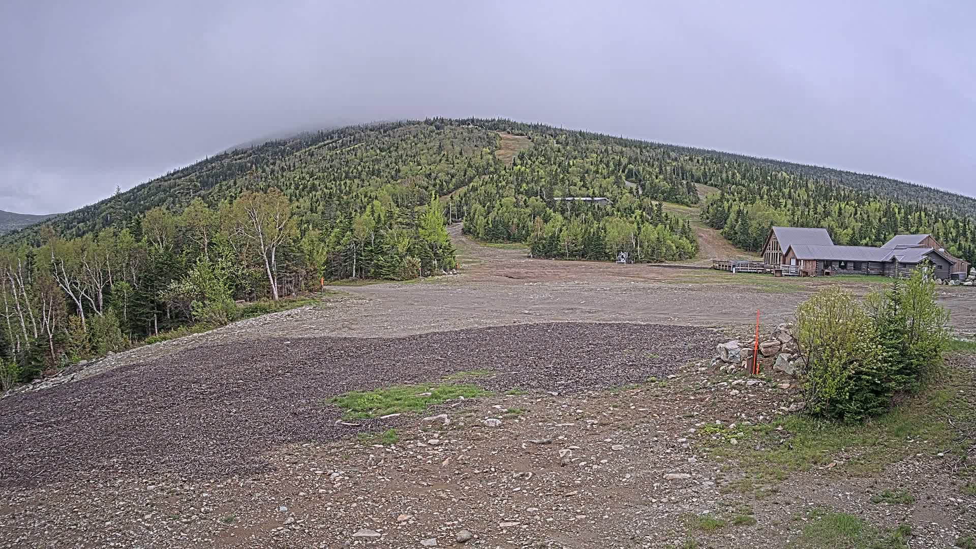 A rocky, mostly barren area at the base of a forested hill under an overcast sky, with a small building complex visible uphill.