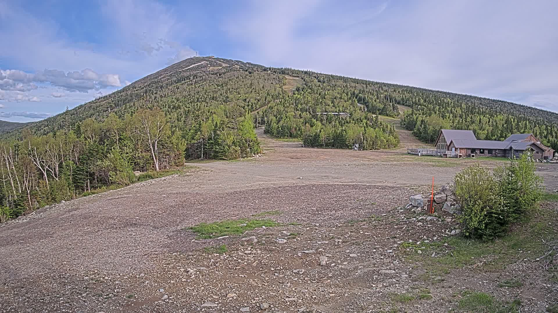 A large, rocky, empty area sits below a forested mountain with a building at its base under a partly cloudy sky.