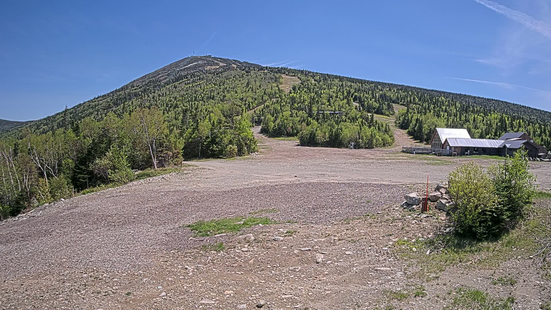 A gravel parking lot sits below a forested mountain with ski runs visible under a clear blue sky.