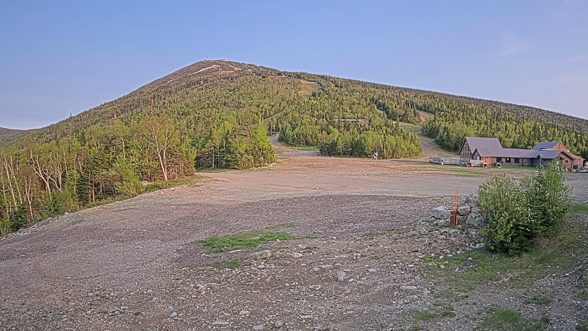 A rocky, gravel area sits in front of a large, tree-covered mountain with a ski slope visible, under a clear, blue sky, and features a wooden building at the base of the mountain.
