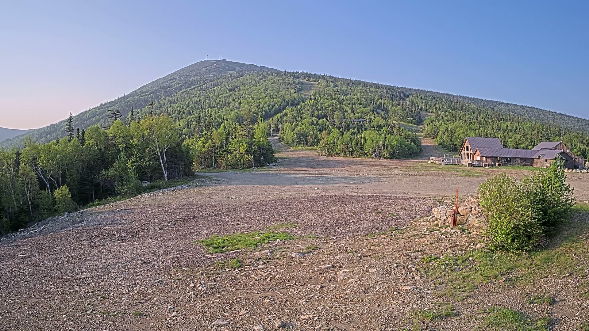 A rocky, empty area sits below a forested mountain on a clear, sunny day, with a wood-framed building visible near the base of the mountain.