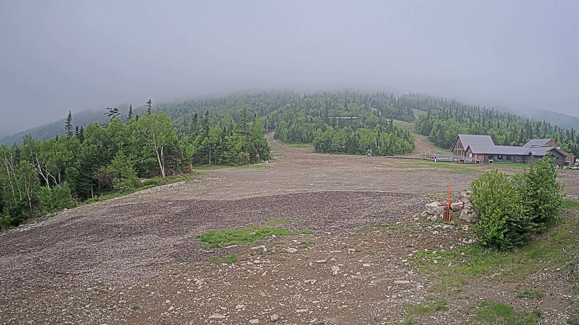 A foggy mountainside shows a rocky, barren ski slope leading to a long, low wooden building nestled among green trees.