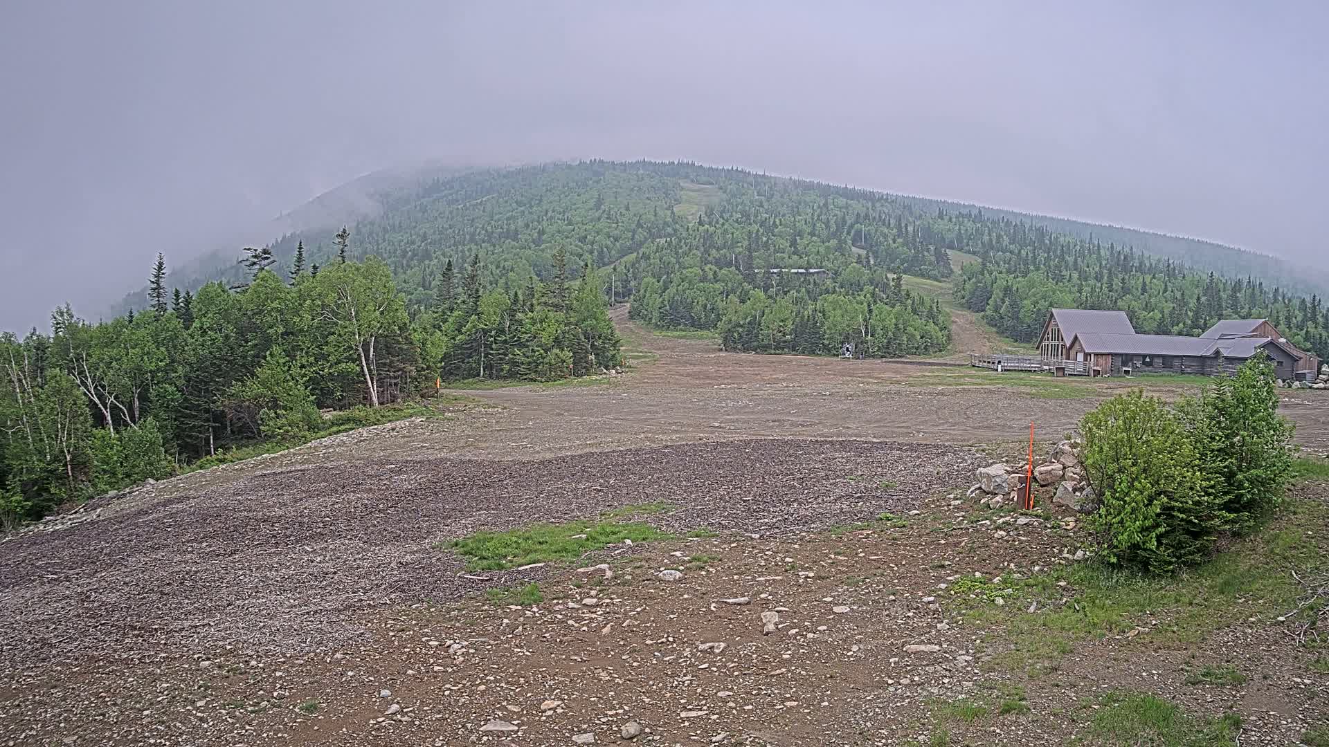 A mostly clear, rocky, flat area sits in front of a forested mountain under a foggy sky, with a building visible in the mid-ground.