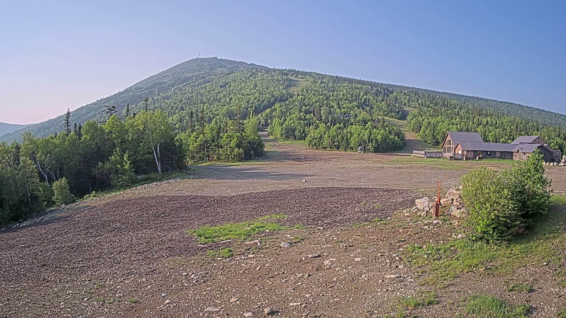 A large, rocky, cleared area sits in front of a ski lodge at the base of a verdant mountain under a clear, blue sky.