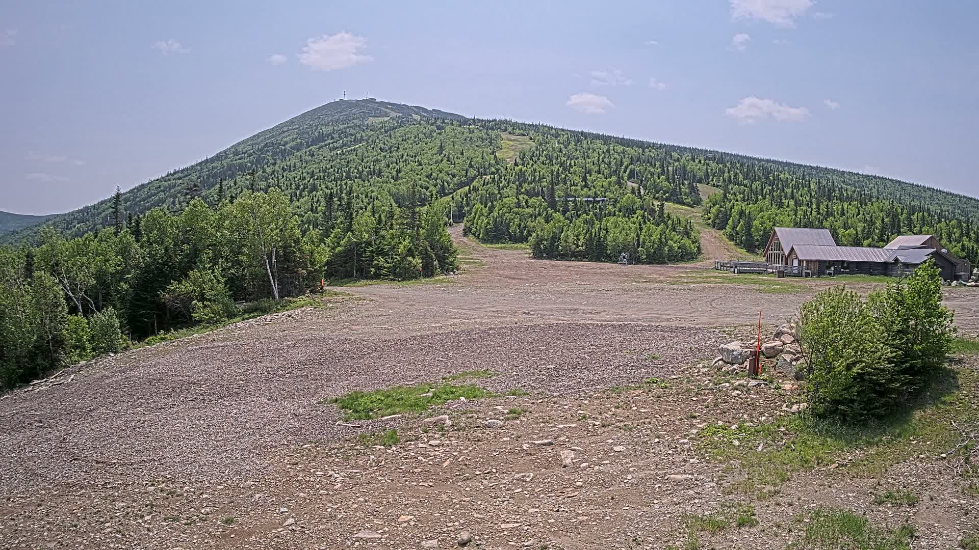 A rocky, empty lot sits in front of a forested mountain with a building complex at its base under a clear, sunny sky.