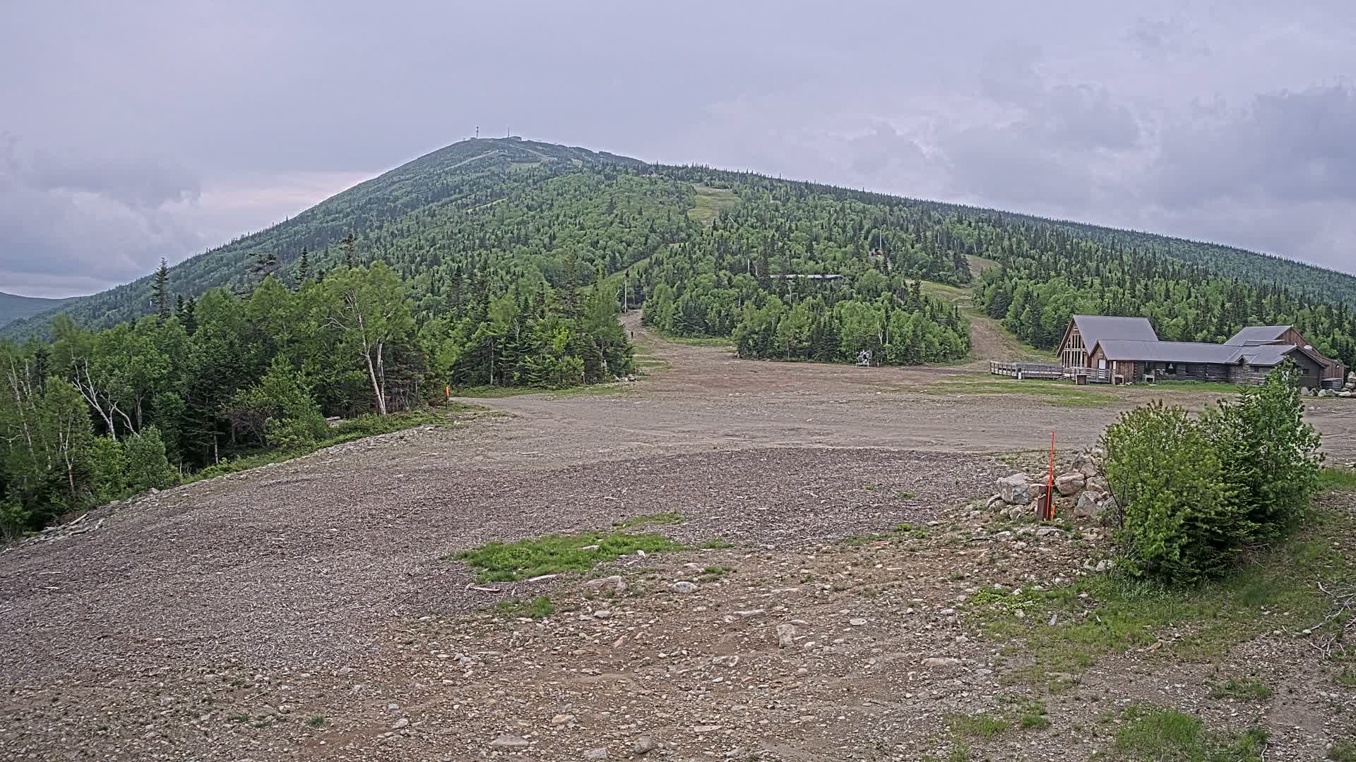 A large, rocky, empty lot sits in front of a ski lodge at the base of a green, tree-covered mountain under an overcast sky.