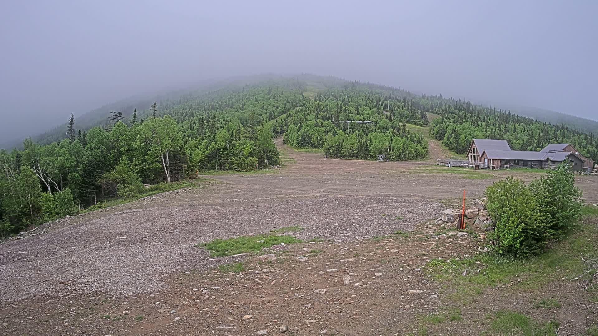 A fog-shrouded mountain slope reveals a sparsely vegetated, rocky clearing with a long building and a forest of green trees.