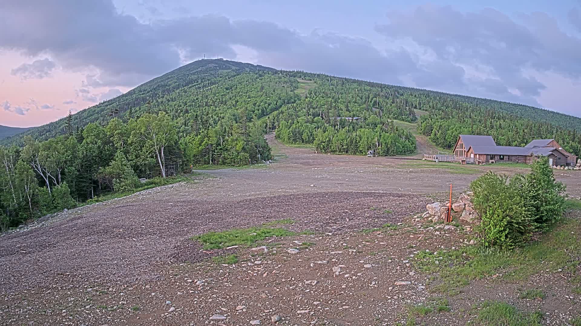 A gravel area sits in front of a ski lodge at the base of a large, green mountain under a partly cloudy, dusky sky.