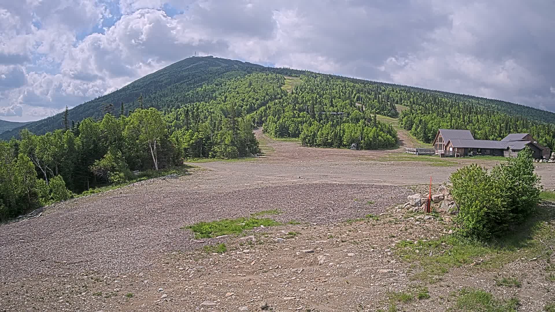A rocky, flat area in front of a ski lodge sits at the base of a large, green mountain under a partly cloudy sky.