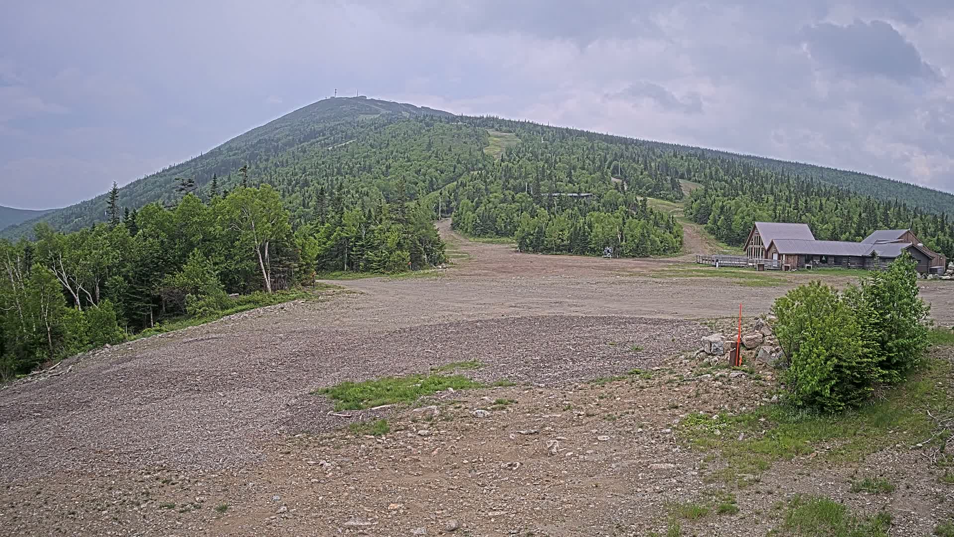 A large, rocky, empty lot sits before a forested mountain under an overcast sky, with a small building complex visible near the base of the mountain.