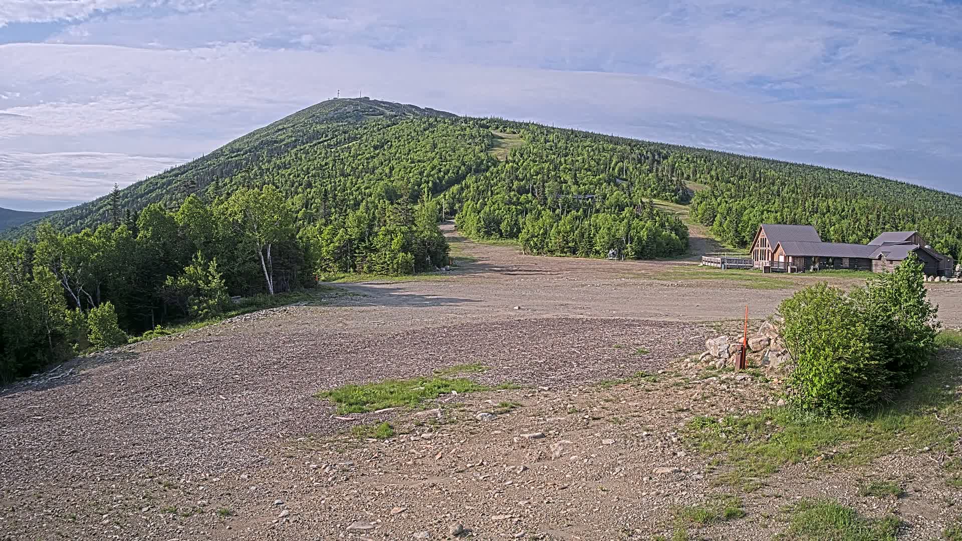A gravel lot sits in front of a ski lodge at the base of a green, forested mountain under a mostly clear, partly cloudy sky.