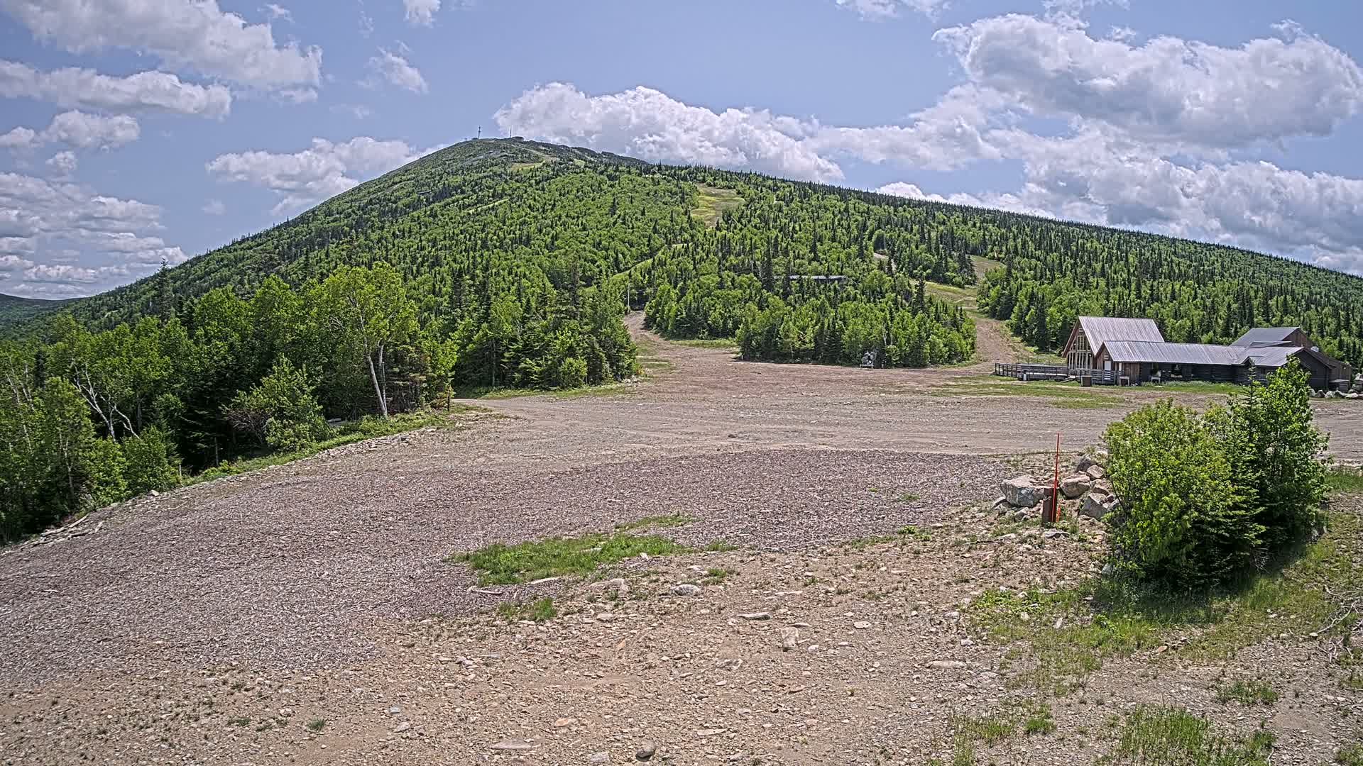 A gravel parking lot sits in front of a few buildings at the base of a verdant, tree-covered mountain under a partly cloudy sky.