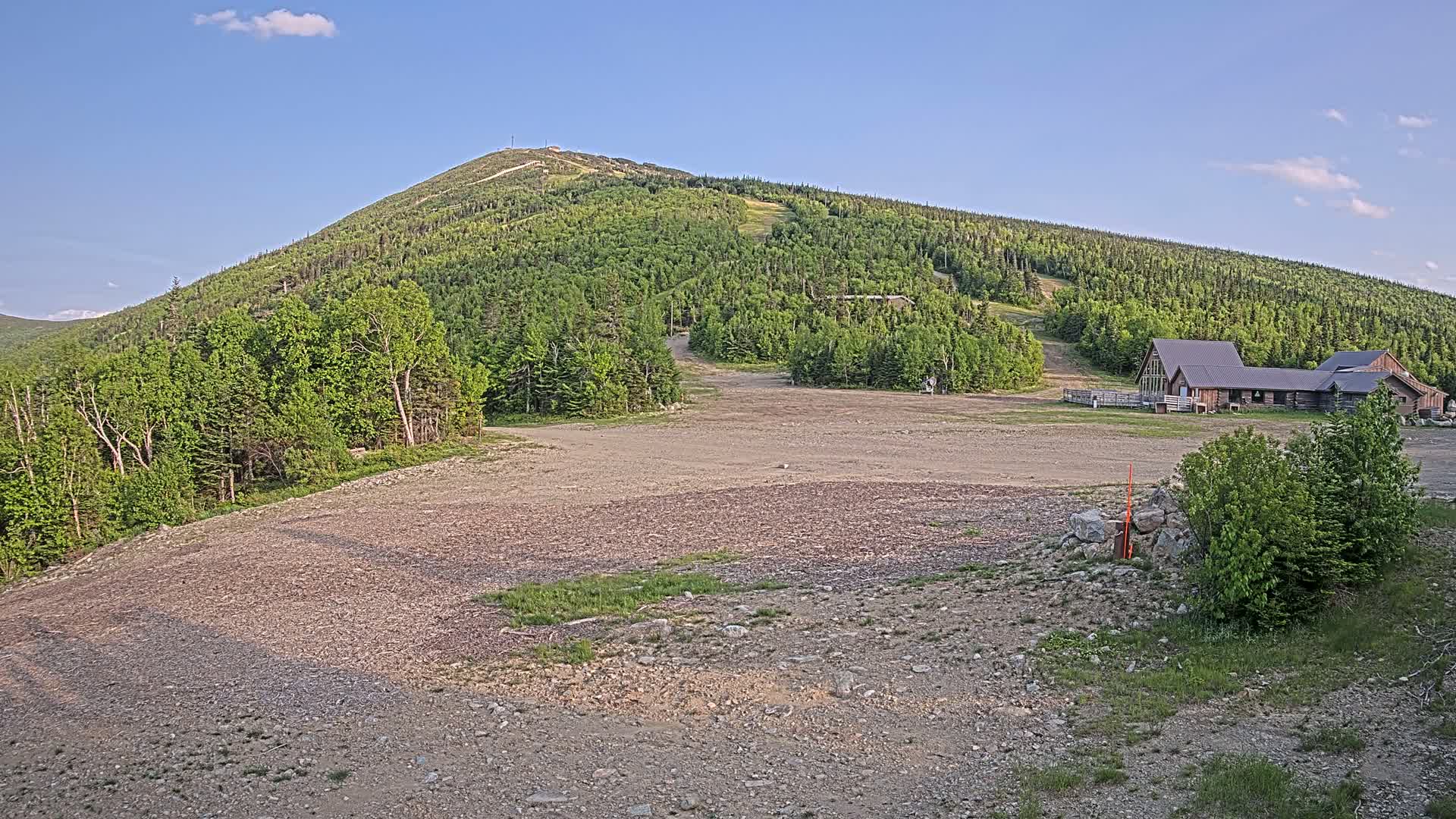 A gravel parking lot sits in front of a few buildings at the base of a verdant, tree-covered mountain under a partly cloudy sky.