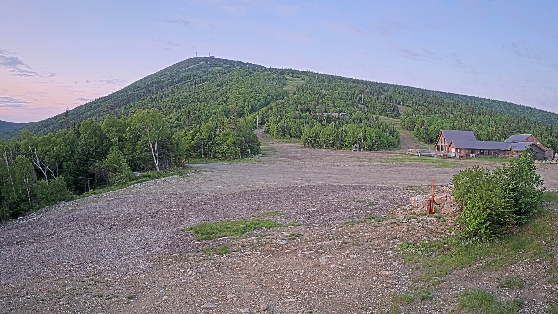 A gravel area sits in front of a ski lodge at the base of a verdant mountain under a twilight sky.