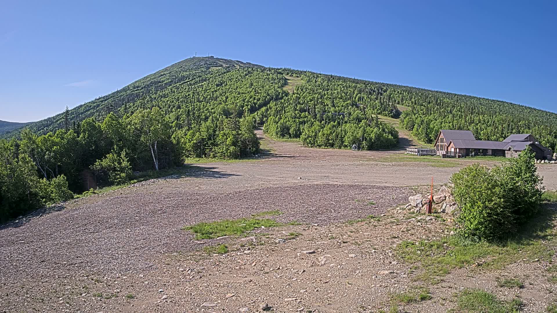 A gravel parking lot sits in front of a ski lodge at the base of a large, green mountain on a clear, sunny day.
