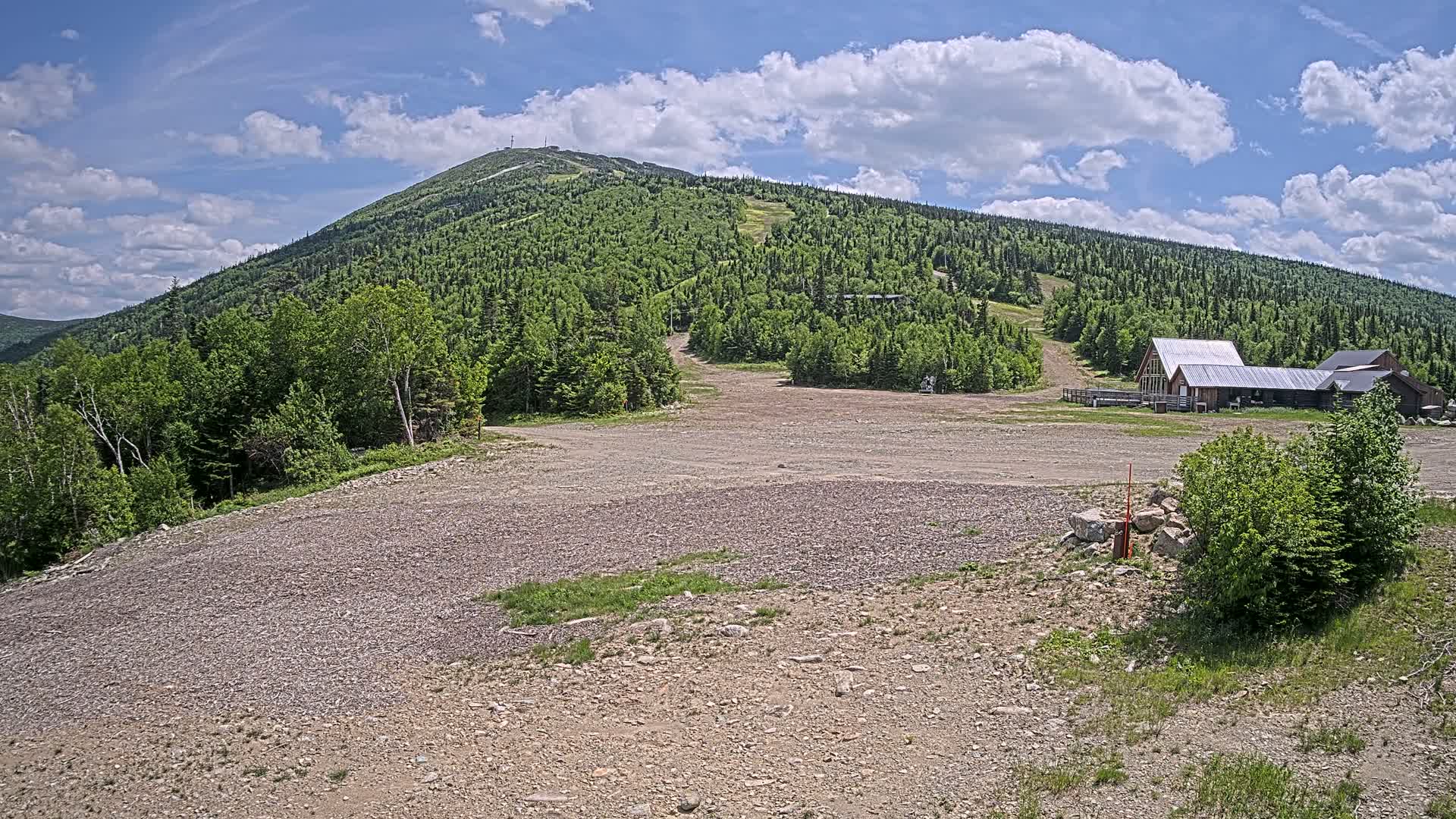 A gravel area sits in front of a ski resort building at the base of a large, green mountain under a partly cloudy sky.