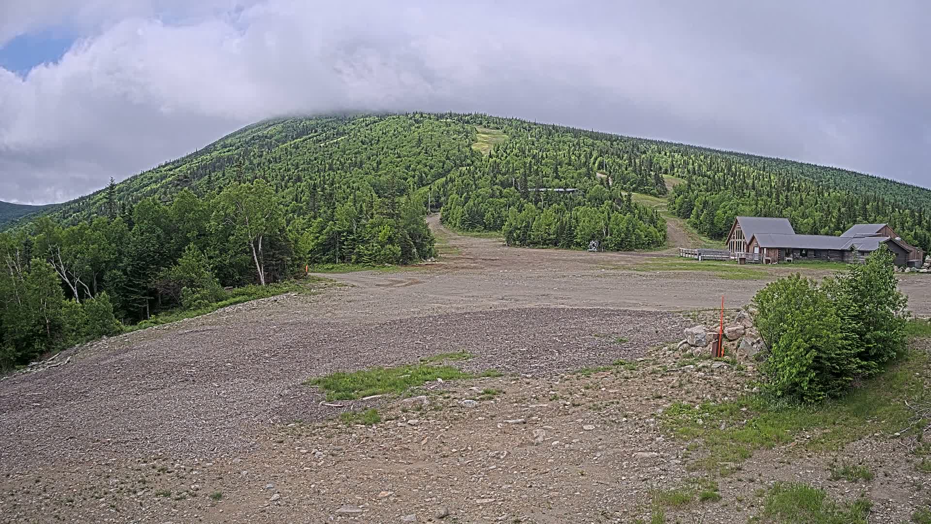 A gravelly area with sparse vegetation sits in front of a forested mountain under a cloudy sky, with a building visible near the base of the mountain.