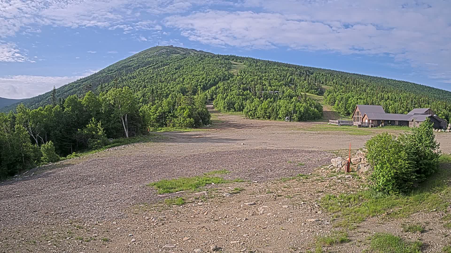 A rocky, mostly barren area sits in front of a large, green mountain with a building at its base under a partly cloudy sky.