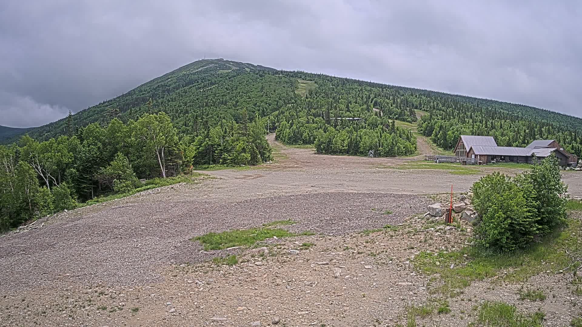 A gravel lot sits in front of a large, forested mountain under an overcast sky, with a few buildings visible near the base of the mountain.