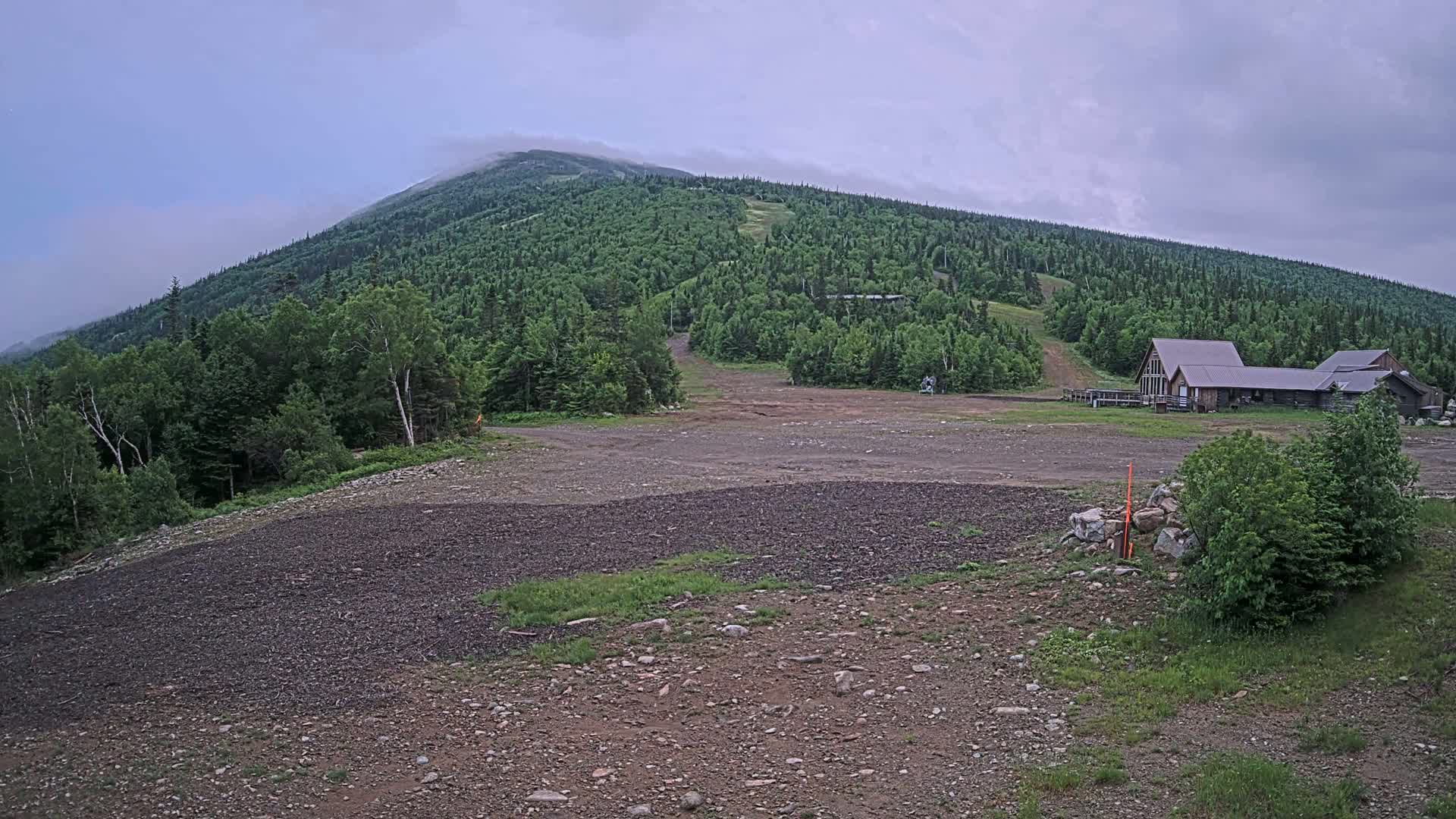 A rocky, mostly barren area sits in front of a forested mountain partially shrouded in fog, with a few buildings visible near the base of the mountain.