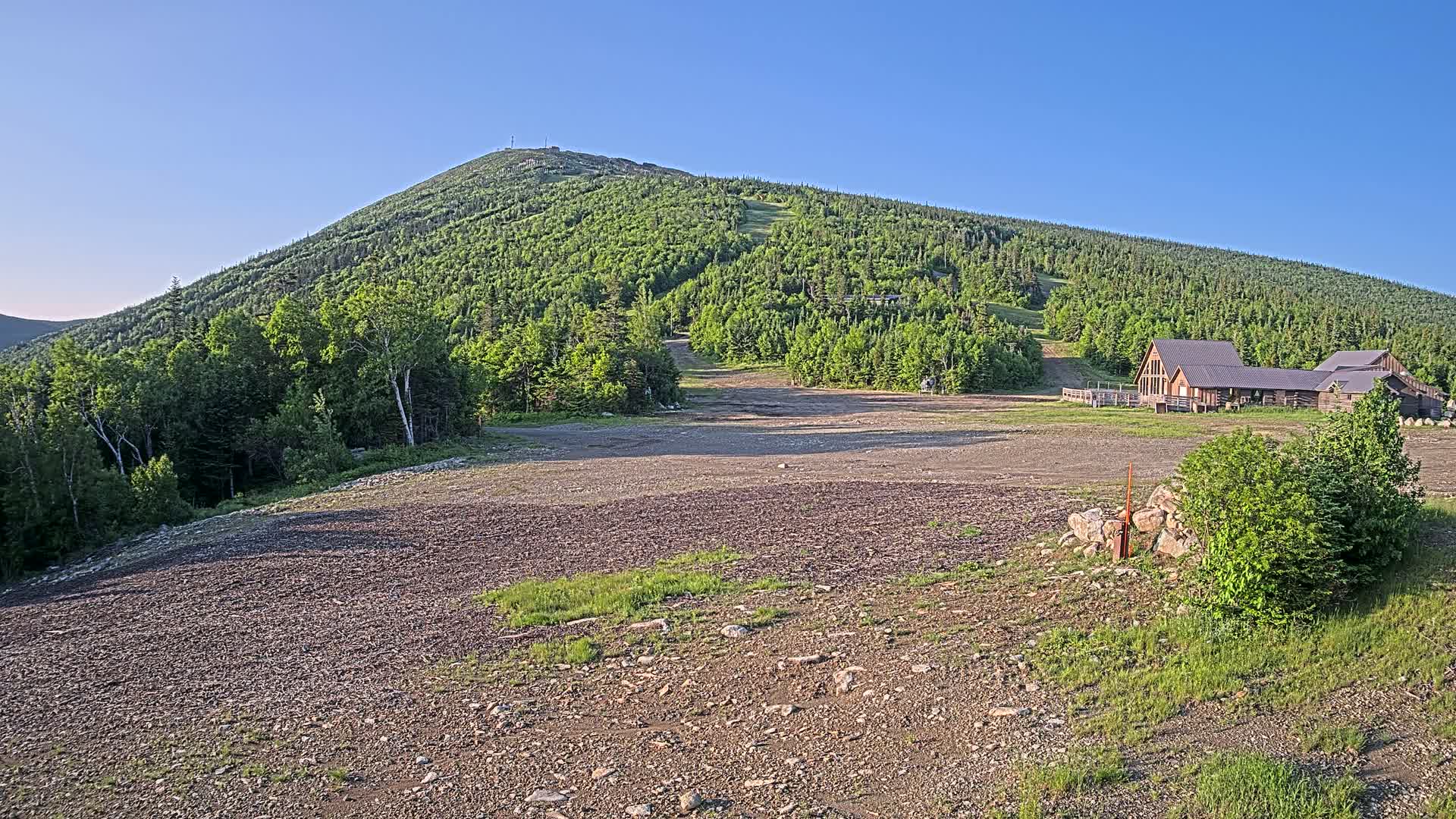 A gravel area sits before a large, green mountain with a building at its base under a clear blue sky.
