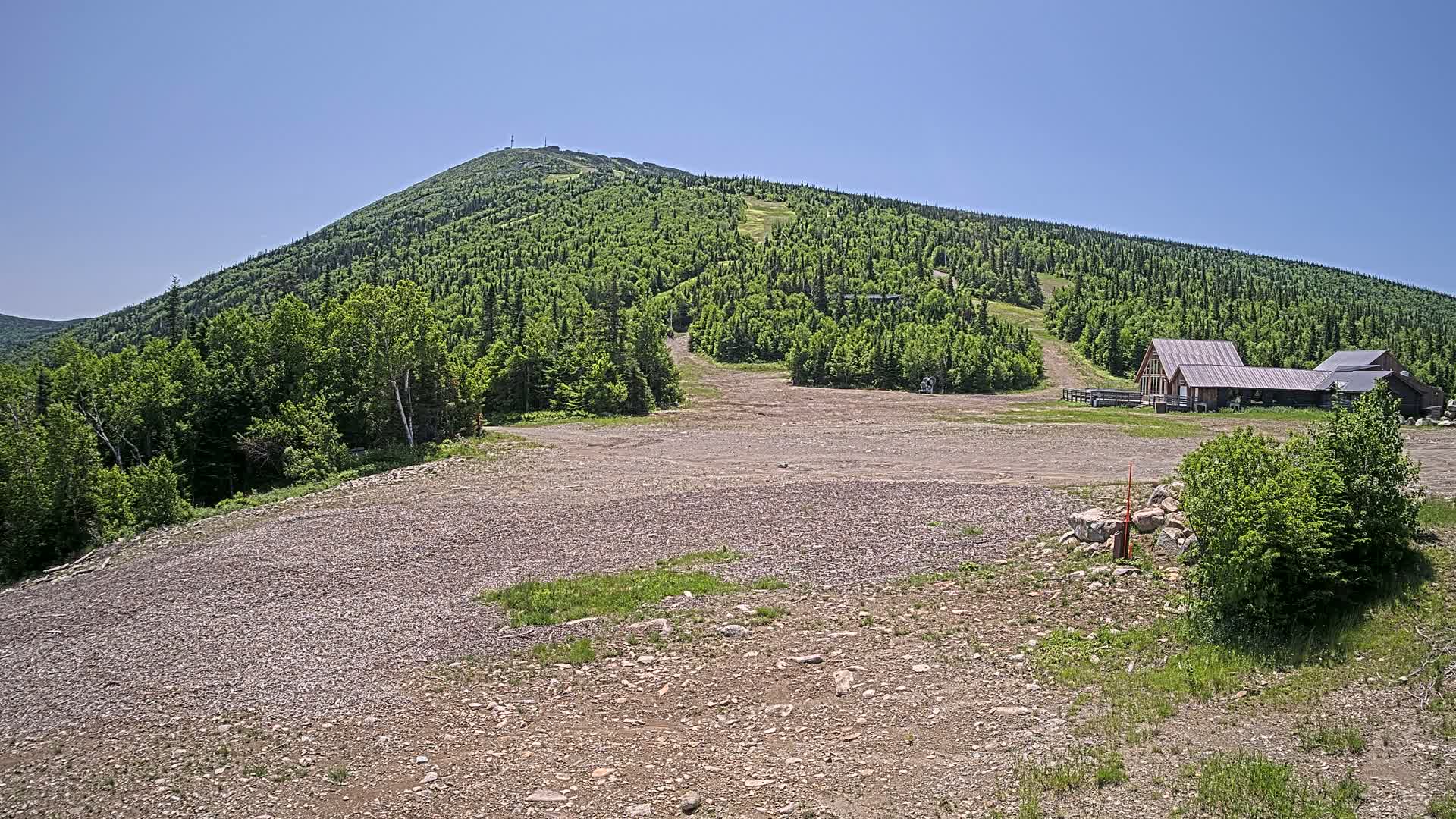 A rocky, grassy area with a small building sits in front of a large, green mountain on a clear, sunny day.